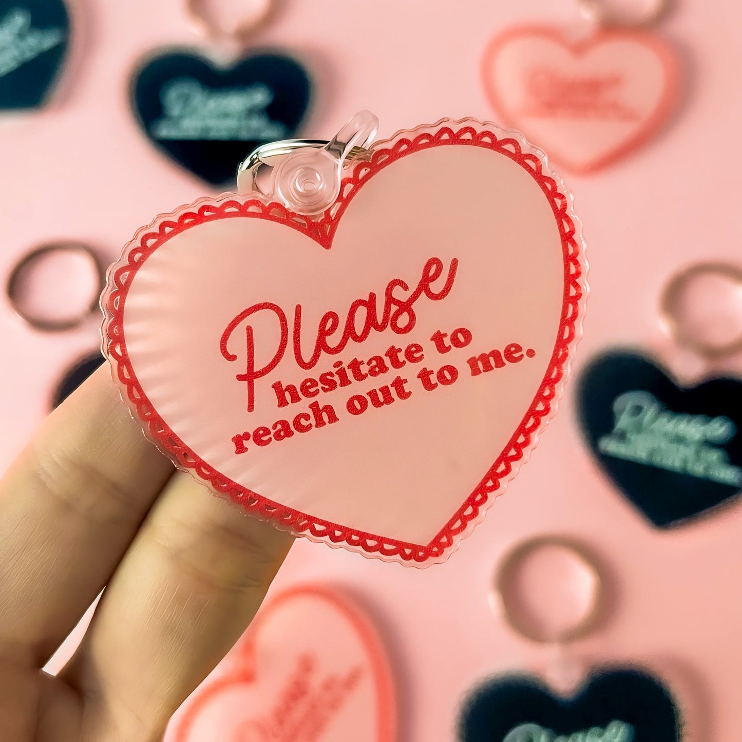 An acrylic heart-shaped keychain, available in either soft pink or bold black, featuring the playful phrase 'Please Hesitate to Reach Out to Me' in elegant white lettering. The keychain is showcased against a neutral background, highlighting its glossy finish and durable construction. It's attached to a classic key ring, ready to add a personal touch to any set of keys or bag