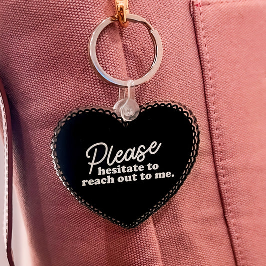 An acrylic heart-shaped keychain, available in either soft pink or bold black, featuring the playful phrase 'Please Hesitate to Reach Out to Me' in elegant white lettering. The keychain is showcased against a neutral background, highlighting its glossy finish and durable construction. It's attached to a classic key ring, ready to add a personal touch to any set of keys or bag