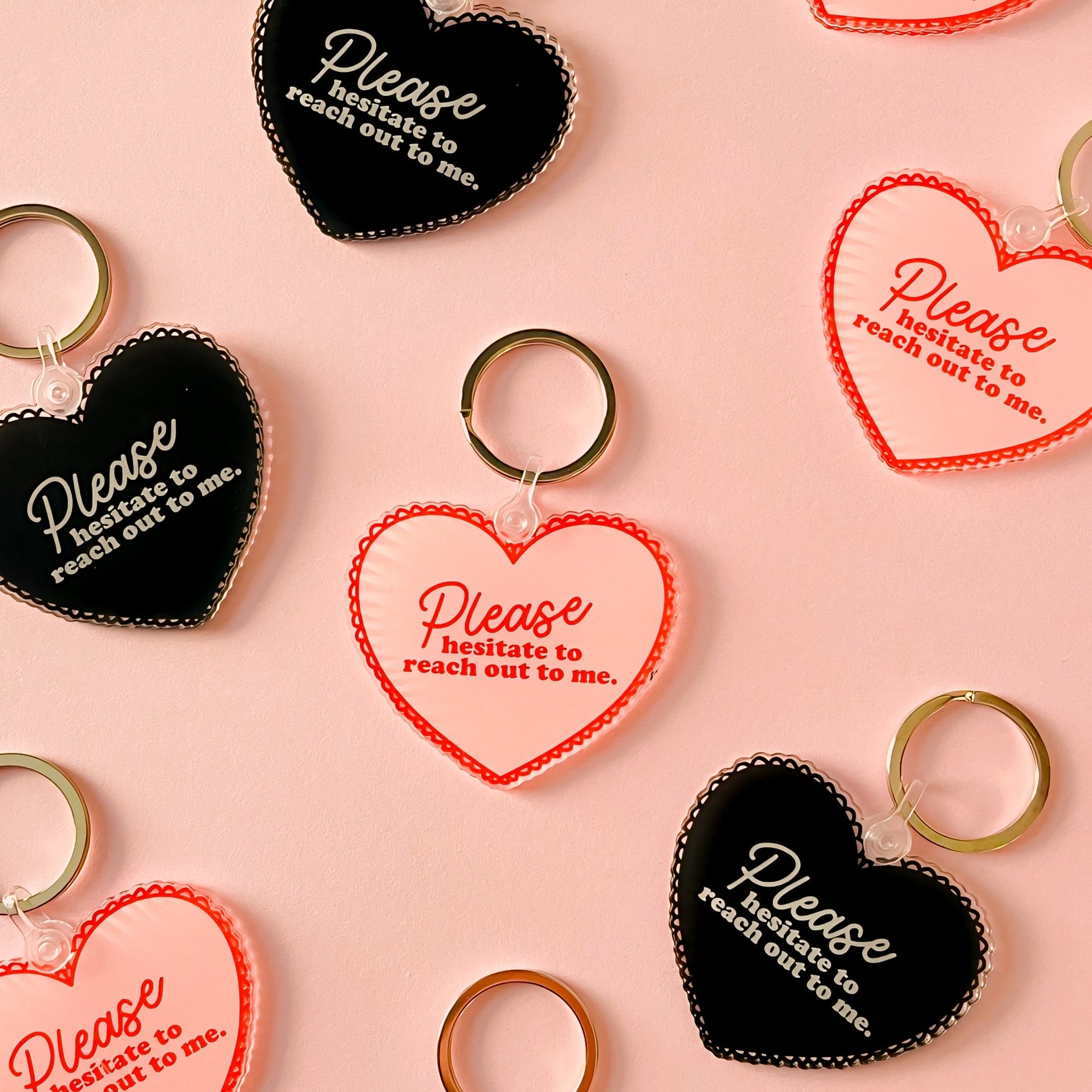 An acrylic heart-shaped keychain, available in either soft pink or bold black, featuring the playful phrase 'Please Hesitate to Reach Out to Me' in elegant white lettering. The keychain is showcased against a neutral background, highlighting its glossy finish and durable construction. It's attached to a classic key ring, ready to add a personal touch to any set of keys or bag